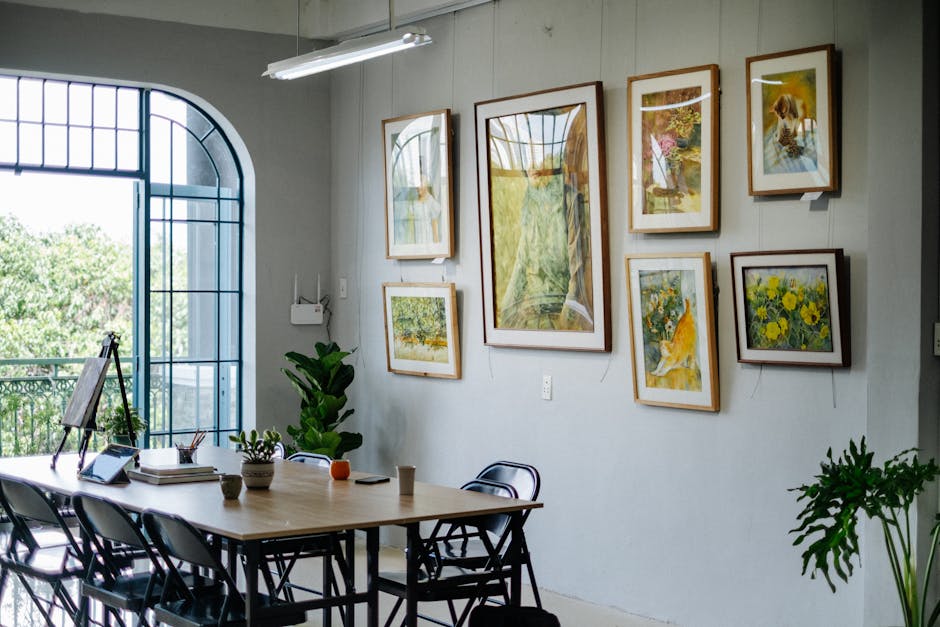 A bright home office featuring framed artwork, greenery, and a large table near a window.