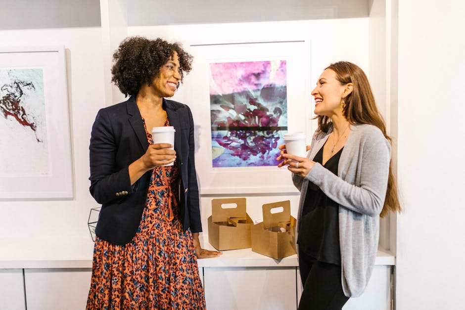Two professional women smiling and chatting during a coffee break in an office art gallery.